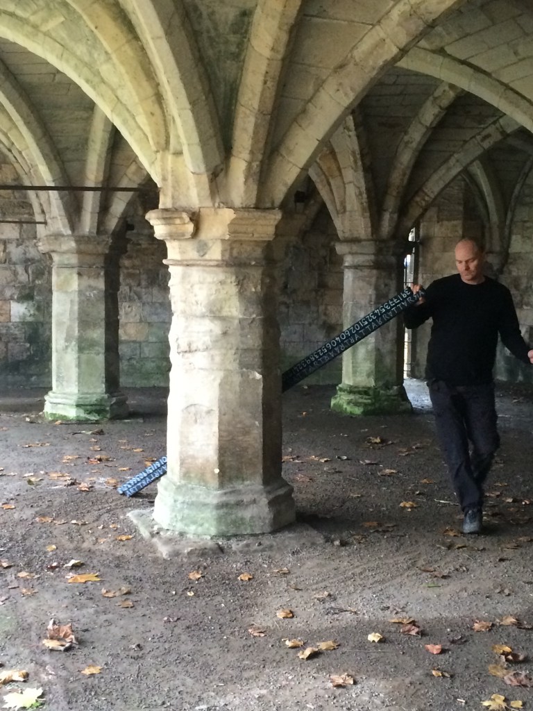  Image of arches, leaves on the ground, a man leans against two thin wooden planks, inscribed with letters and numbers.