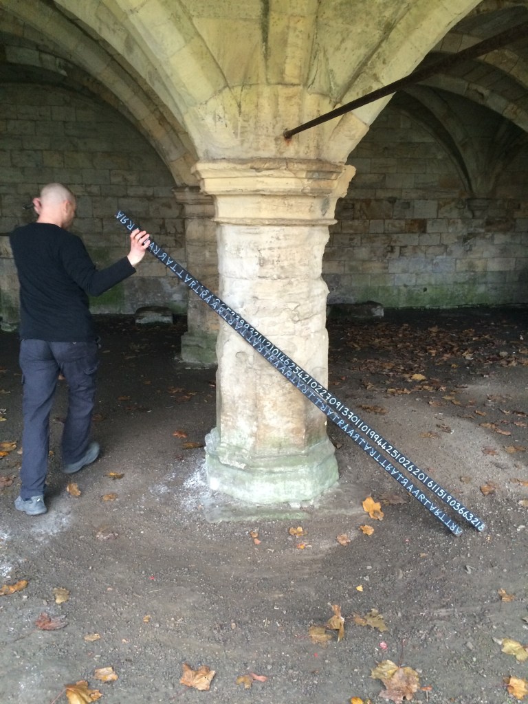 Image of arches, leaves on the ground, a man faces away from the camera, dragging  the two thin wooden planks, inscribed with letters and numbers.