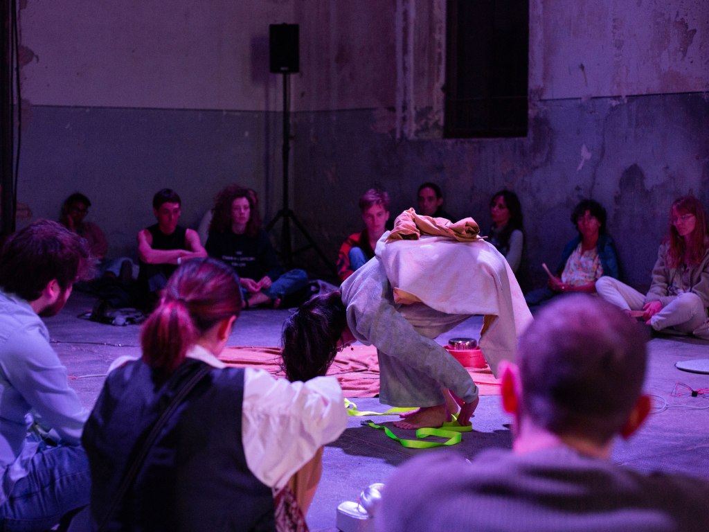 a performer at the centre of the room is bent over double. There black hair covers their face. They are wearing grey clothes with a pink blanket draped over their back. They are surrounded on all sides by seated audience members