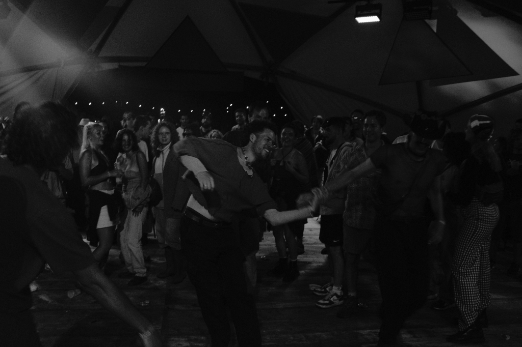 a black-and-white image of many people dancing inside a domed tent. In the foreground are two men holding hands and pulling slightly away from each other. The man on the left wears a T-shirt that is pulled up by his movement and a chunky necklace, and is smiling. The man on the right wears a mesh T-shirt and a cowboy hat. There are many people around them watching their movements with joy on their faces