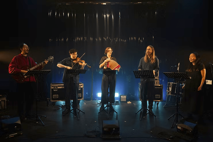 Five people on stage. Each has a music stand in front of them, the three on the left are playing instruments (a guitar, a violin and an accordion). The lighting is sharp on their faces but otherwise muted.