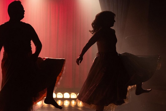 two silhouetted figures wearing swishy skirts perform a cancan kick in front of a red curtain and golden lights