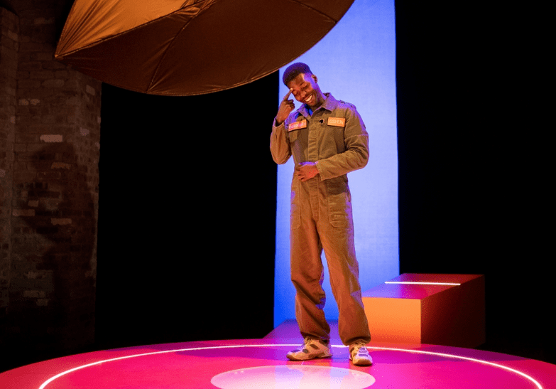 A dark skinned man with cropped hair stands on  circular stage lit pink and orange, wearing pale brown overalls. Above him is a fabric shape, behind him are bands of black and blue, and a brick wall to his right. He is pointing at his forehead and smiling