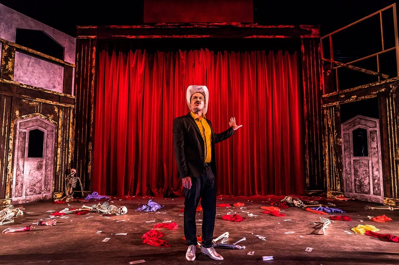 A man with glasses, a moustache and dark hair, wearing a dark suit, a yellow shirt and shiny sneakers, stands at the front of the stage, one hand gesturing to the red curtain behind him. The stage is strewn with torn clothing and skeletons, On either side of the stage are theatre flats painted with doorways, distressed to look down at heel.