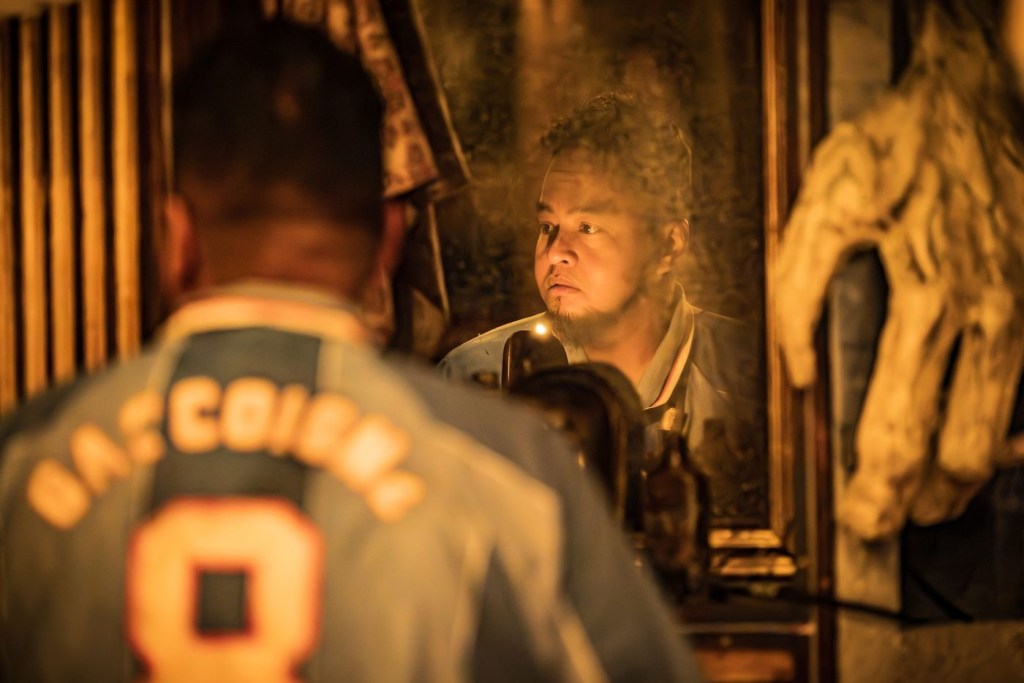 A man with pale brown skin, short dark hair and a beard that doesn't cover his chin stands in front of a mirror, looking to his left. His reflection is sharp, and he has a thoughtful expression on his face. Although it's blurred, 