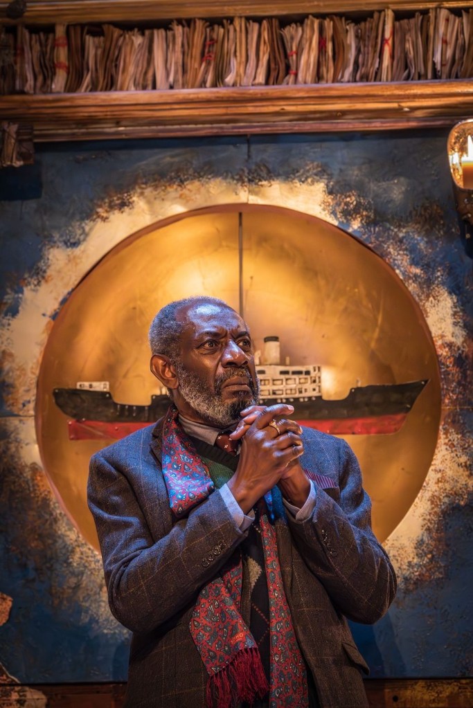 A man with dark skin and salt-and-pepper hair stands in front of a sculpture of a ship, which hangs in front of a gold circle like a sun. Above the sun is a row of papers bound together. The man wears a woollen suit, a green jumper, a brown tie and a burgundy paisley silk scarf, has a serious expression, and has his hands clasped as if in prayer.