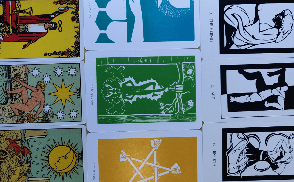 Nine tarot cards from different eras arranged in a square within a pill-shape. At the left of the square are images of a magician, a star above a naked person pouring water into a pond, and two dogs howling at the moon. Down the centre of the square are blue, green and yellow cards depicting cups, a person hanging by the ankle from a tree, and five crossed swords. At the right of the square are three black and white cards depicting stylised figures kneeling or posed in movement.